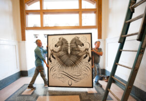 Two men carrying a large framed artwork featuring two stylized horses facing each other, surrounded by intricate swirling lines, in a grand indoor space with marble floors and large windows.