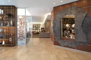 Interior view of a modern lounge area featuring a decorative wooden wall with shelves and sculptures, a glass door leading to a dining space, and a large rustic circular opening into a coffee bar with modern fixtures in the background.