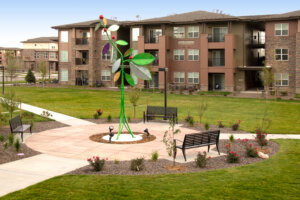 A landscaped courtyard featuring a colorful, large flower sculpture at the center, surrounded by circular pathways, benches, and flower beds, with modern apartment buildings in the background under a clear sky.