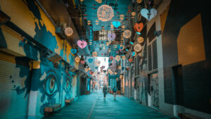 A vibrant alleyway decorated with colorful hanging hearts and spherical light fixtures, featuring a mural of large eyes on the left wall. Two individuals walk towards the camera along a path lined with benches.