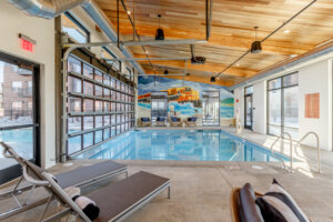Indoor swimming pool area featuring a mural of a train over waves, large glass doors allowing natural light, and several lounge chairs with striped cushions.